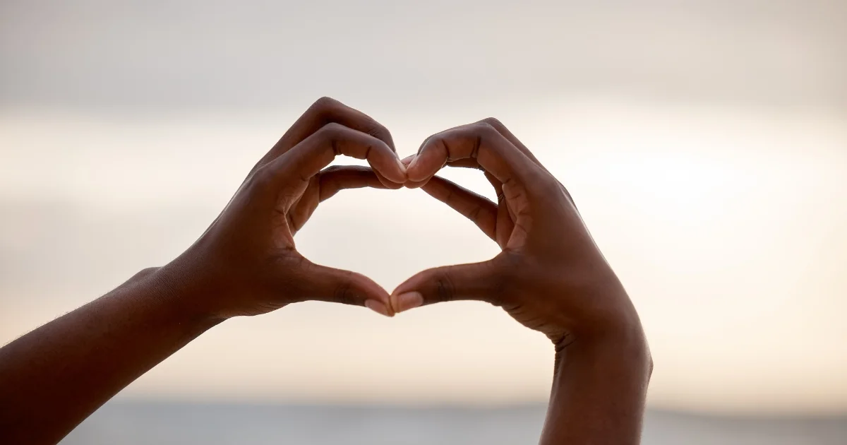 Hands forming a heart shape against a soft, cloudy sky background.